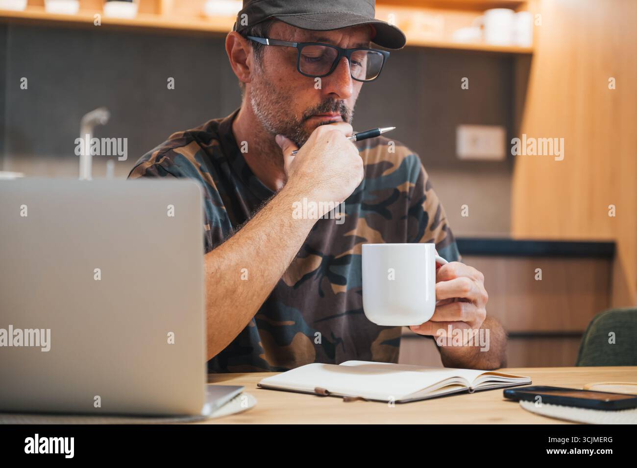 Man wearing army hat and military camouflage shirt drinking coffee and making to do list in the morning in modern indoor setting, selective focus Stock Photo