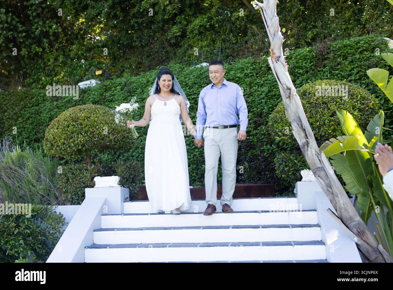 Walking down garden steps, bride and groom holding hands smiling joyfully Stock Photo