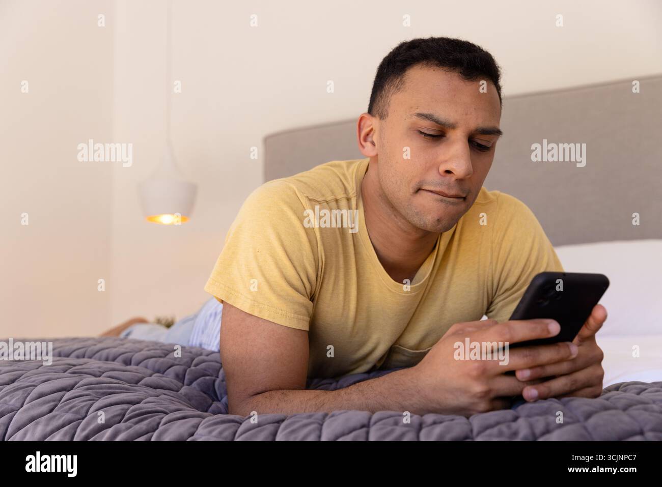 Young man lying on bed using smartphone, focused on screen Stock Photo