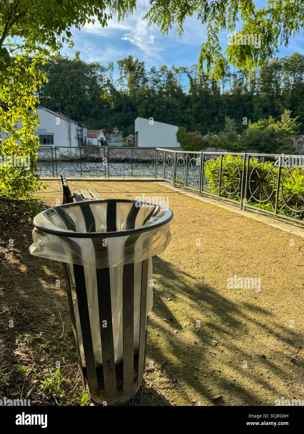 Riverside park scene with a stripped trash bin, wooden banch, metal fence, and canal view on a sunny day. - Smartphone Captured Stock Image