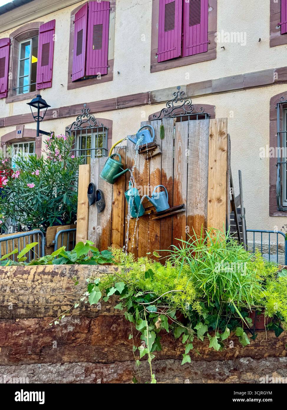 artistic outdoor fountain made from metal watering cans on a wooden panel, in front of a building with purple shutters and flowers. - Smartphone Captured Stock Image