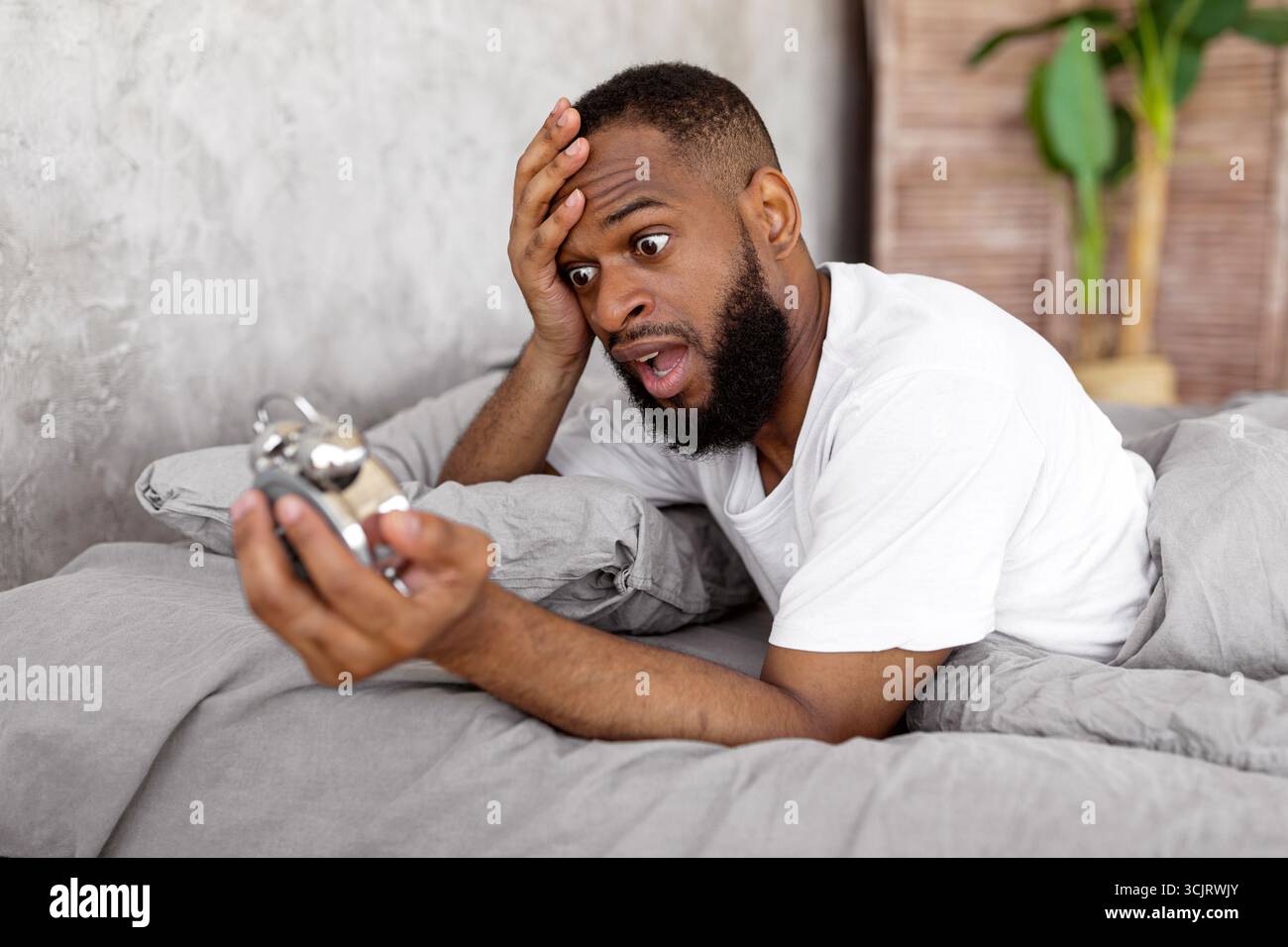 Shocked black man looking at clock in bed Stock Photo