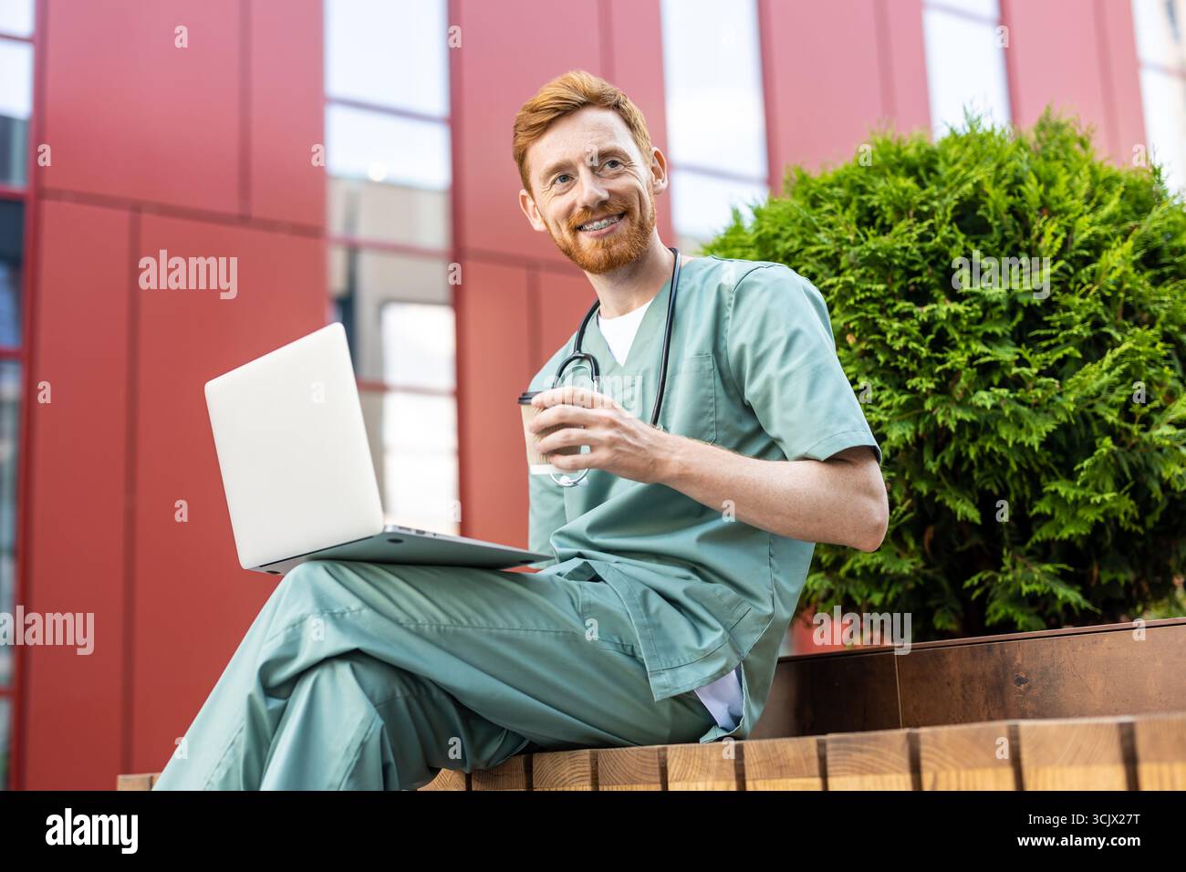 Smiling man in scrubs holding takeaway drink while using laptop Stock Photo