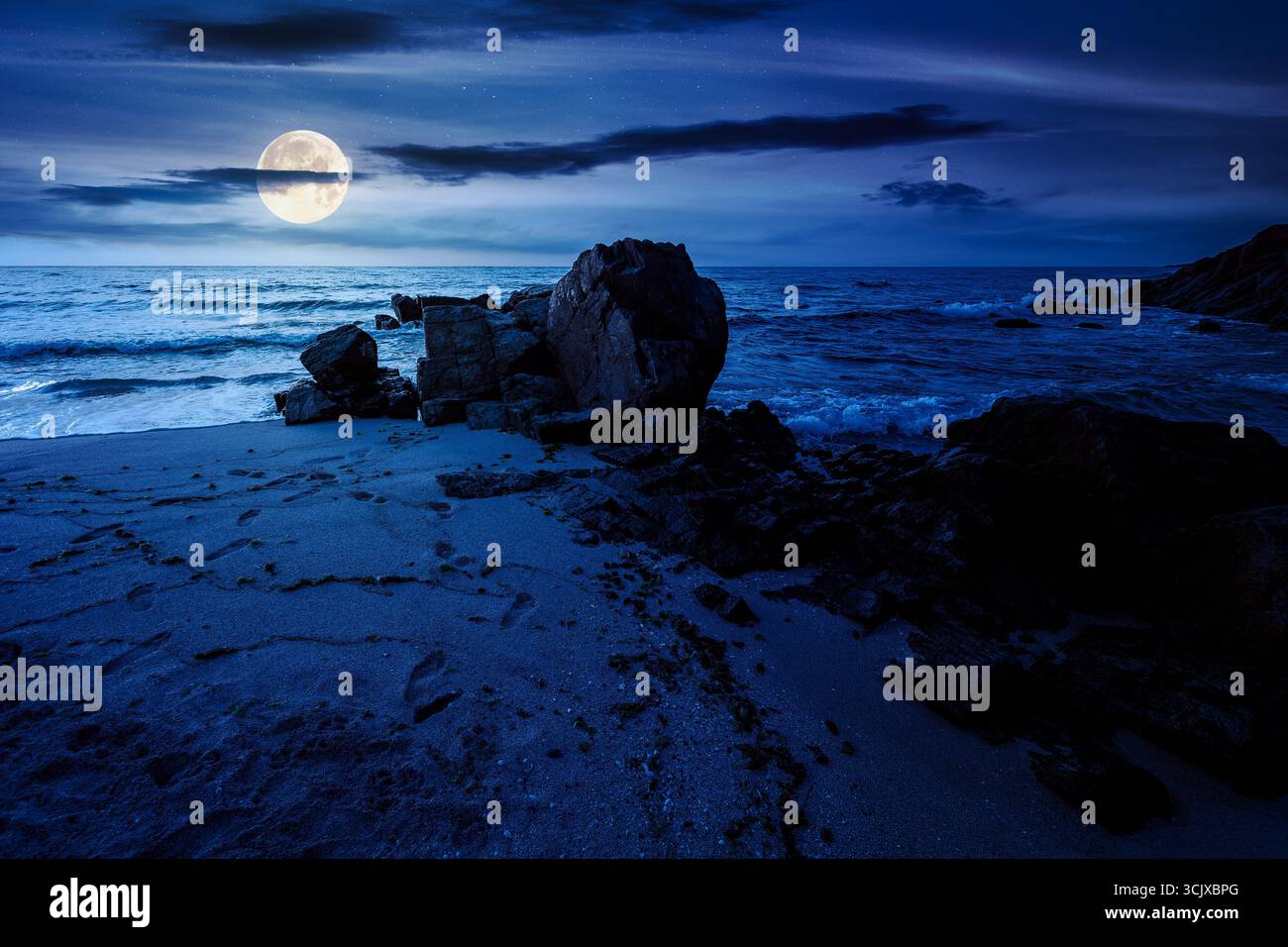 landscape by the sea at night in summer. beautiful beach with rocks washed by calm waves in full moon light. view to horizon under dark sky with cloud Stock Photo