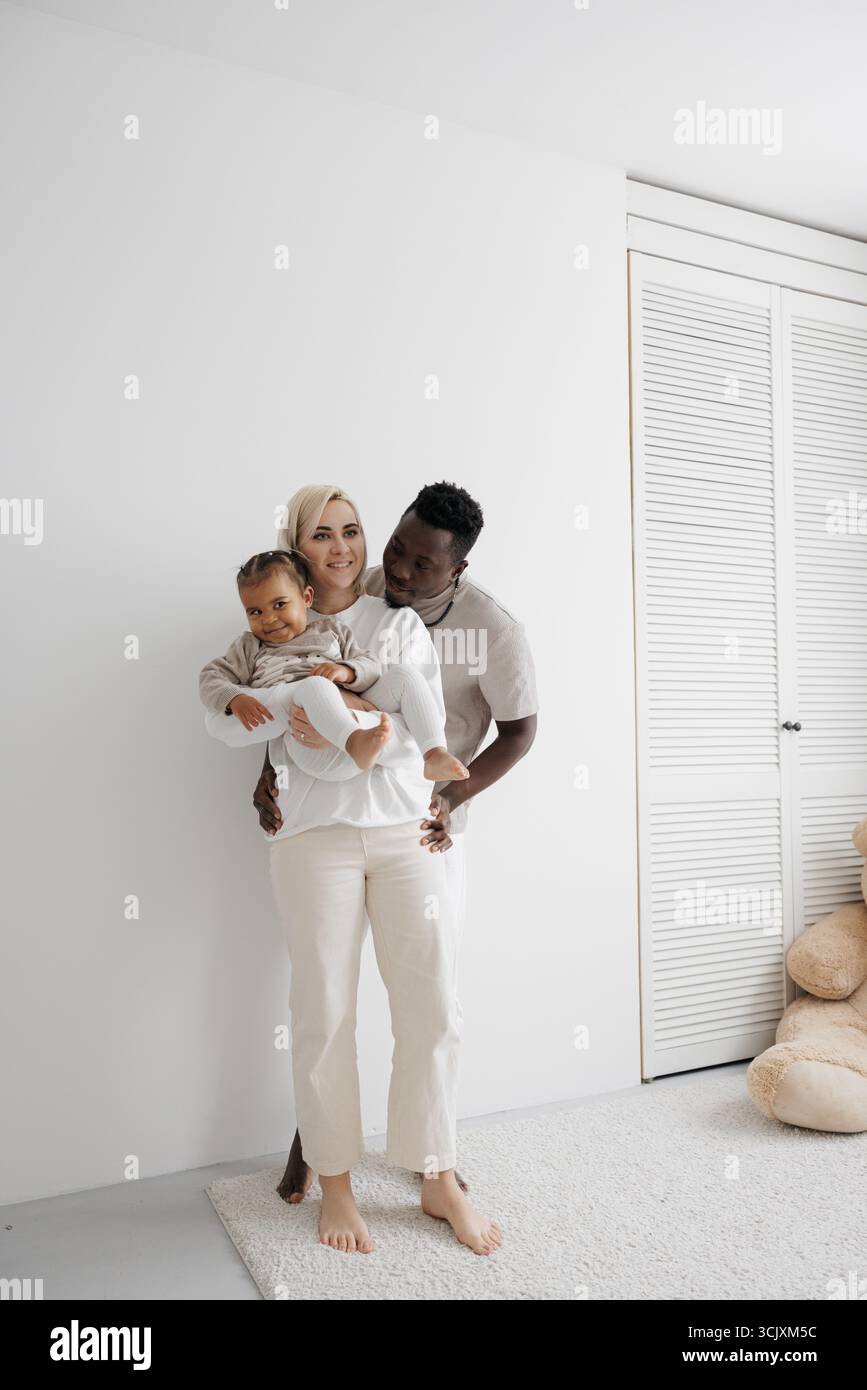 Loving biracial family holding their daughter and embracing against a white wall. Concept of interracial family. Stock Photo