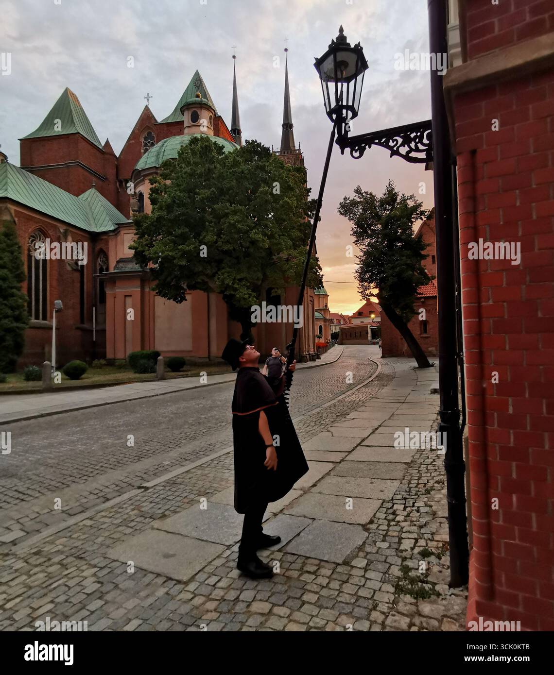 Wrocław lamplighter in hat and cloak lighting gas lamps on Cathedral Island at dusk—historic tradition, Poland, tourism, street scene, Ostrów Tumski. - Smartphone Captured Stock Image