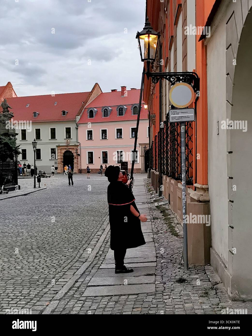 Wrocław lamplighter in hat and cloak lighting gas lamps on Cathedral Island at dusk—historic tradition, Poland, tourism, street scene, Ostrów Tumski. - Smartphone Captured Stock Image