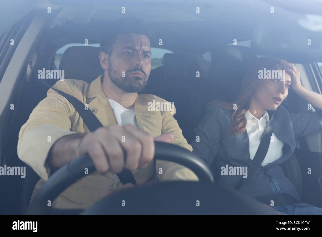 Man in drivers seat with annoyed expression gripping steering wheel, while upset woman in passenger seat holds her head. Tense atmosphere after an arg Stock Photo