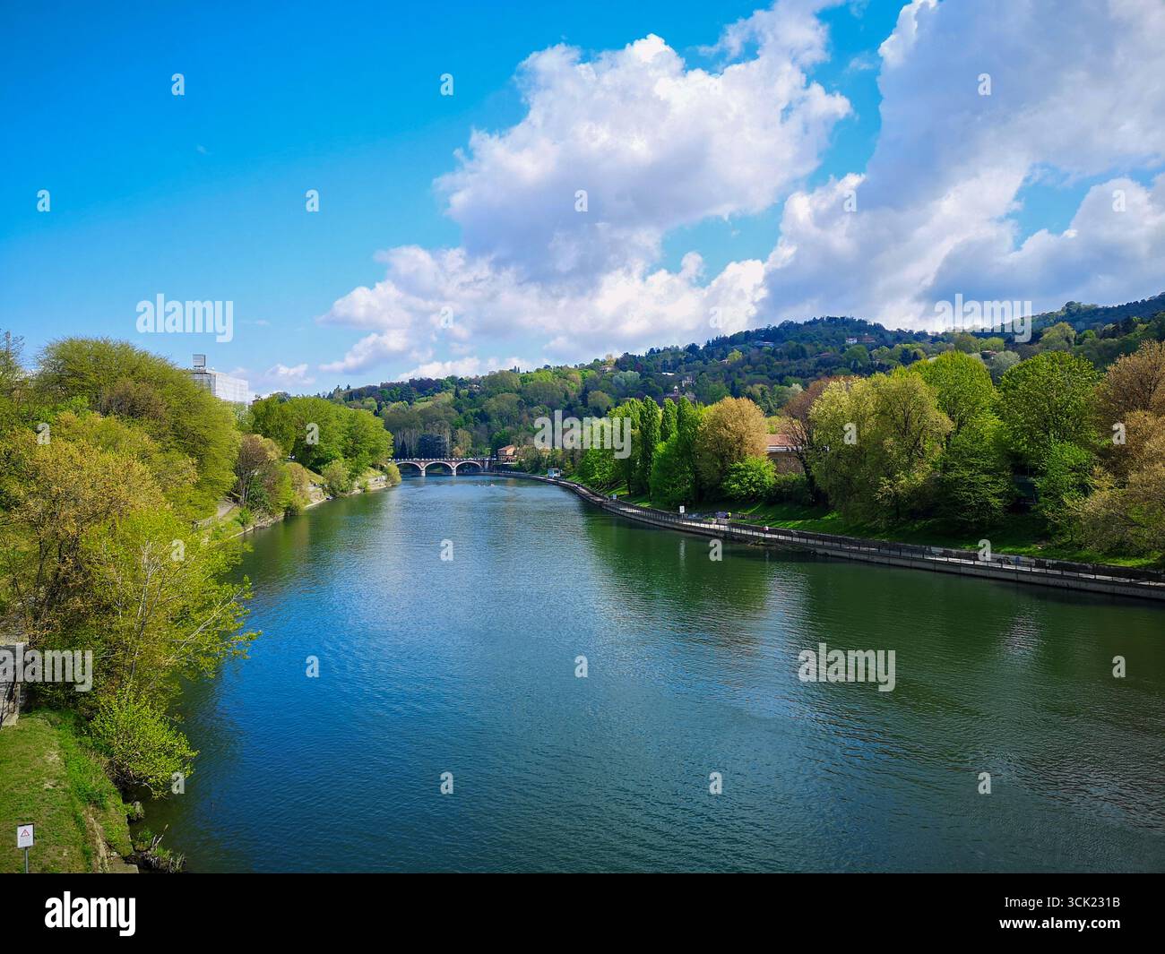A scenic view of the Po River flowing through Turin, Italy, on a bright, sunny day. The calm, blue water of the river is bordered by lush green foliag - Smartphone Captured Stock Image
