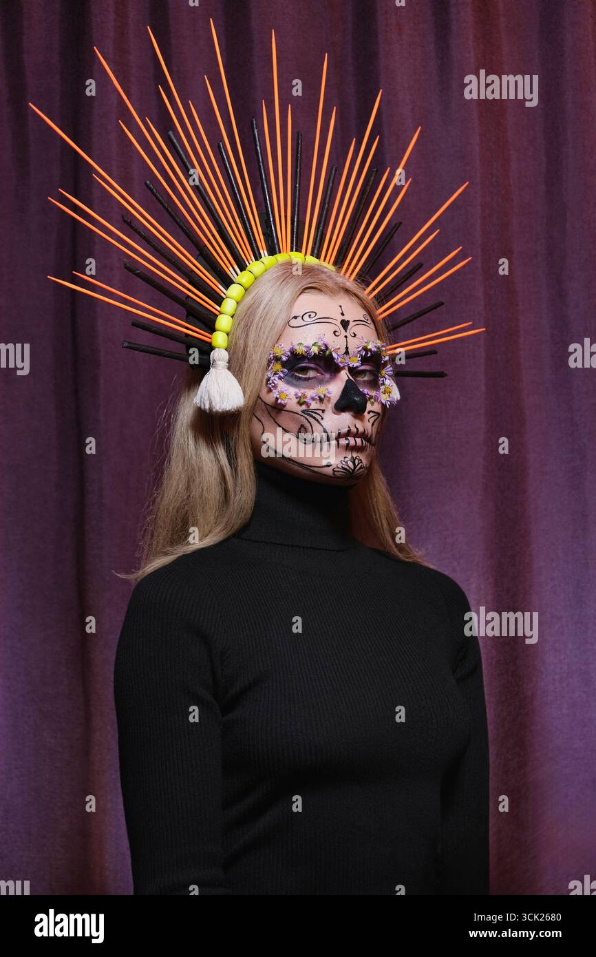 Blonde woman with sugar skull makeup and large orange crown shaped like sun rays. Unique ceremonial look reflecting cultural heritage, beauty Stock Photo