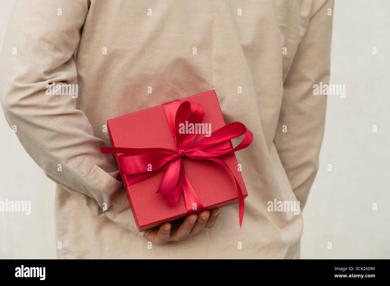 Man hiding red gift box tied with ribbon behind their back, wearing beige shirt for surprise girlfriend. Birthday present suggests surprise or special Stock Photo