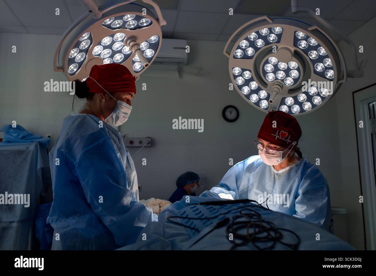 Two medical professionals perform a surgical procedure in a well-equipped operating room. They are dressed in sterile blue gowns and red caps, illumin Stock Photo
