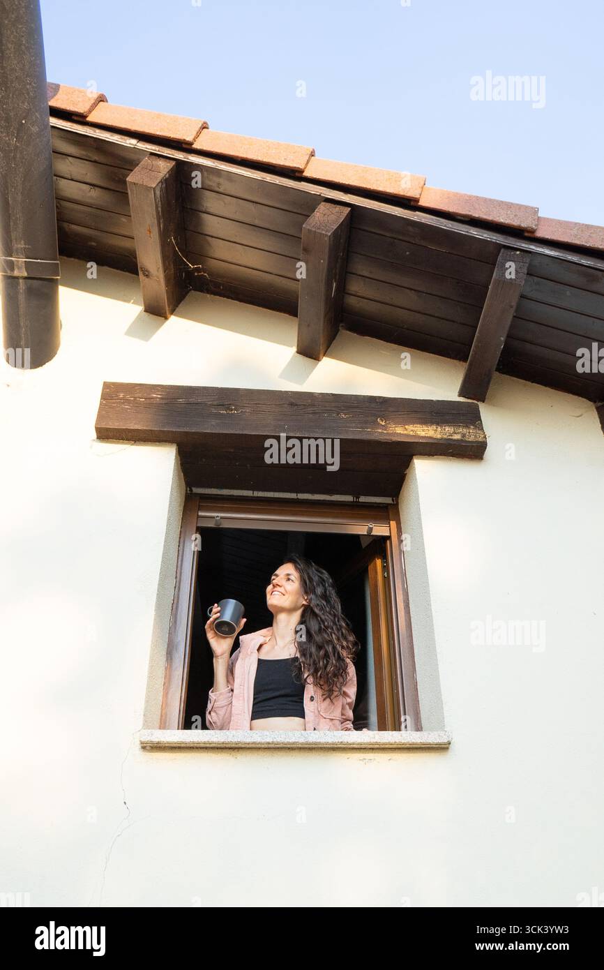 Smiling woman drinking coffee and looking outside the window of her house Stock Photo