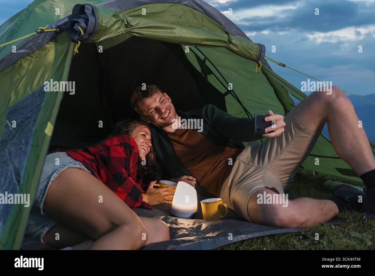 In a serene outdoor setting, a couple relaxes inside a tent, sharing laughs while taking a selfie with a soft light and warm drinks. Stock Photo