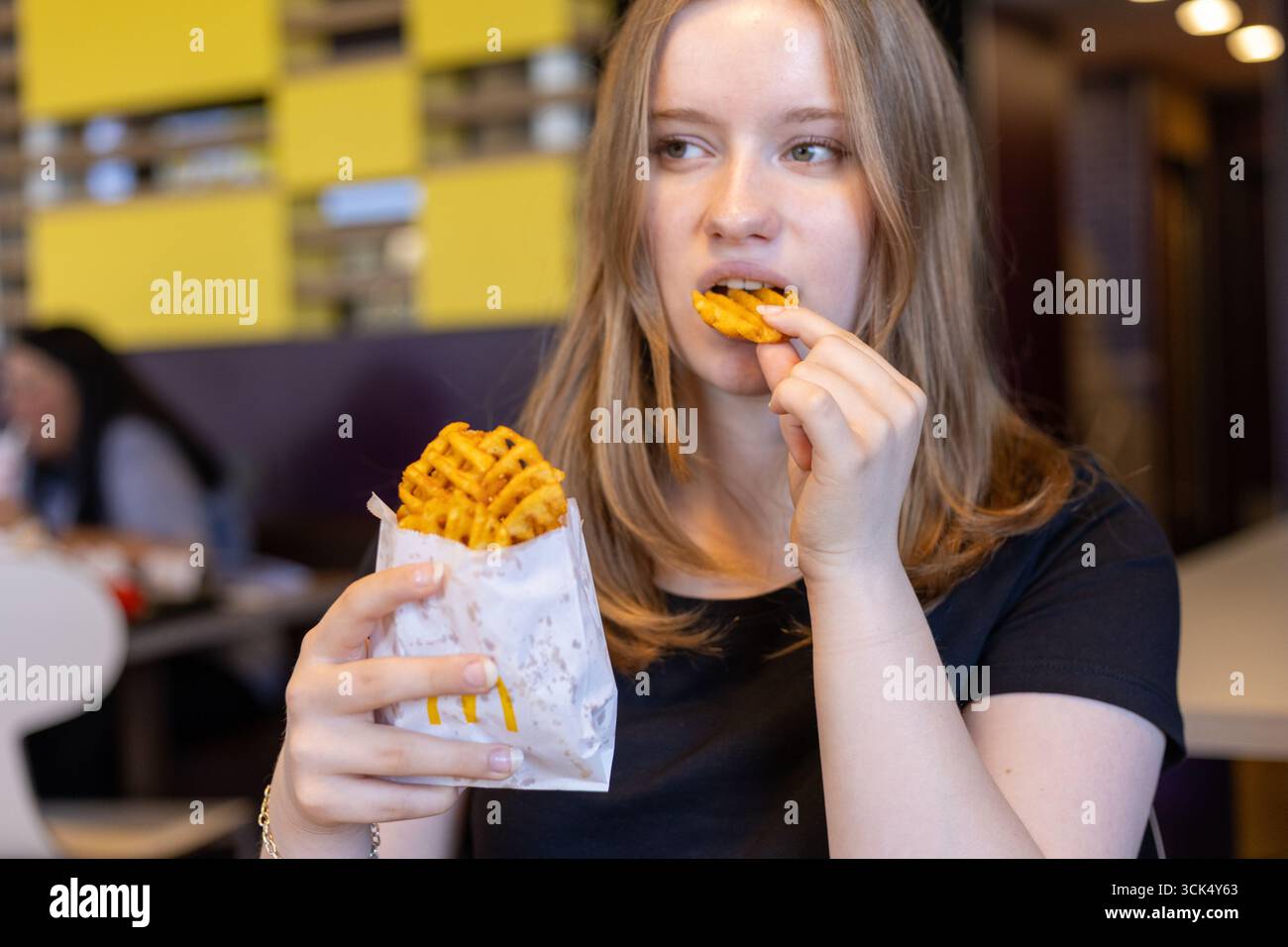 Kerpen, Germany - August, 9, 2025: A young woman takes a bite of waffle fries at a vibrant in McDonalds, modern eatery. She enjoys a casual lunch whil Stock Photo
