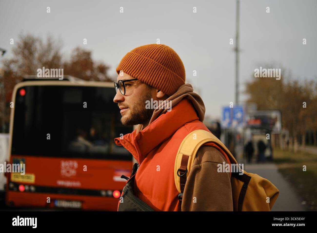 Side profile of a man in an orange beanie and glasses standing near a bus, dressed in a red vest and brown hoodie with a yellow backpack. Stock Photo