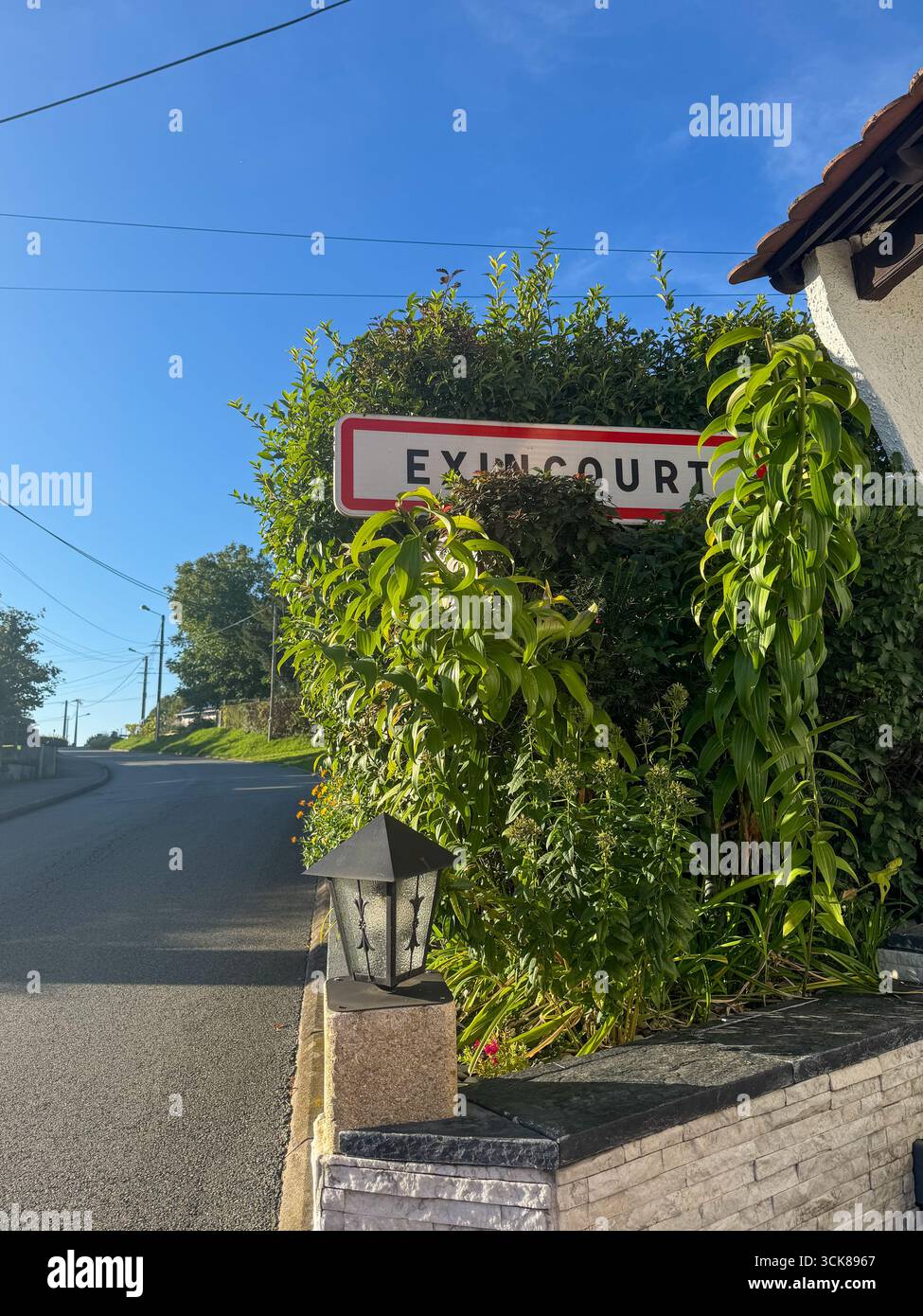 Quiet street in Exincourt, France, with a curving road, stone wall, town sign, charming houses, small gardens, lush greenery and sunny blue sky - Smartphone Captured Stock Image
