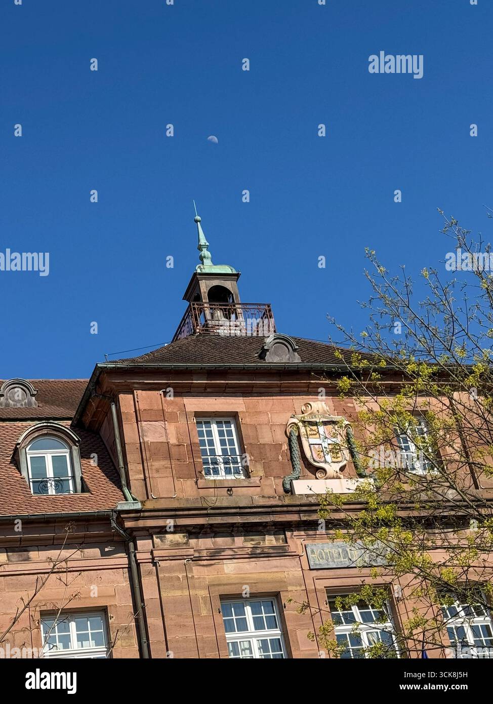Historic building in Place Cuvier Monbeliard, France, with steep roof, tower, spire, reddish-brown stone facade, decorative crest, with the moon - Smartphone Captured Stock Image