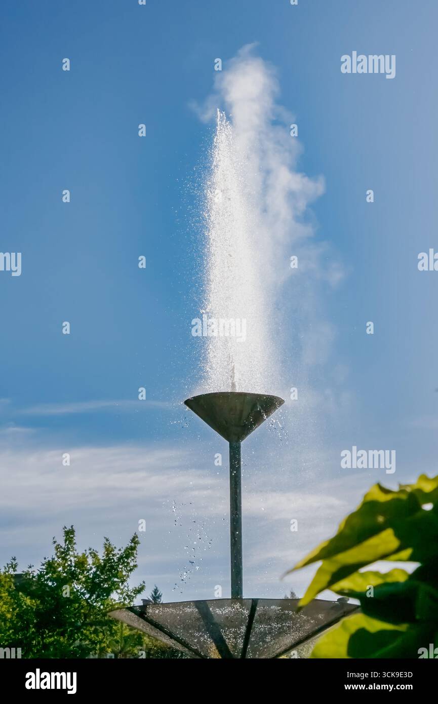 A vertical fountain shoots a tall jet of water into the air, creating mist and droplets under bright sunlight, photographed against a clear blue sky i Stock Photo