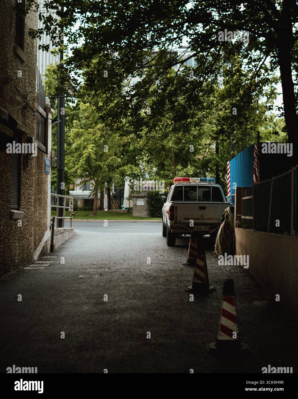 Parked patrol vehicle in a shaded urban alley, flanked by lush trees and safety cones— a serene snapshot of neighborhood watch in the city Stock Photo