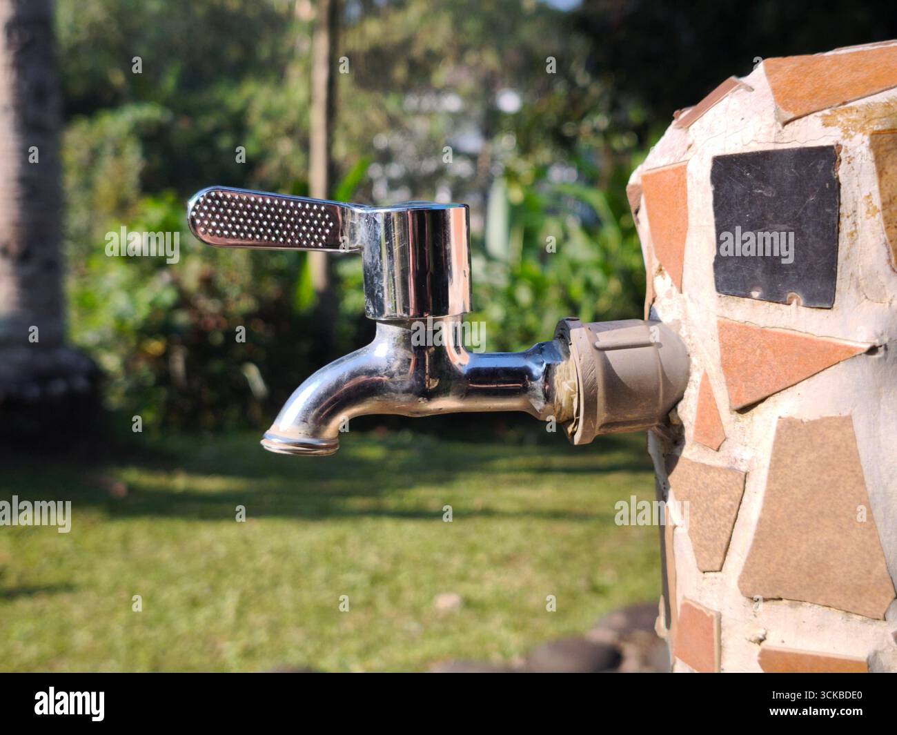 A shiny outdoor water faucet attached to a wall decorated with ceramic tiles. The background shows a garden with green grass and plants Stock Photo