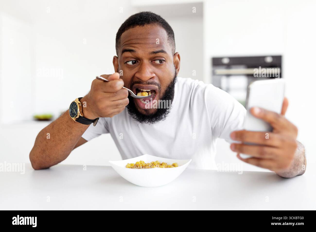 Man Enjoying Breakfast While Using Smartphone in Modern Kitchen Stock Photo