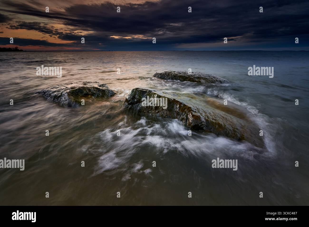 Wave-washed rocks stand out in the sea under a dramatic, dark evening ...
