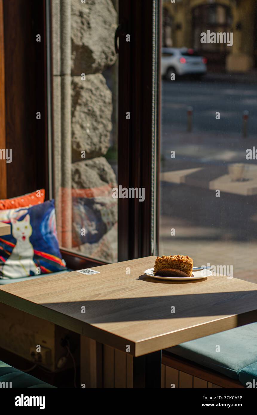 Slice of carrot cake on wooden cafe table by window with colorful decorative pillows and sunlight, cozy urban coffee shop interior. Stock Photo