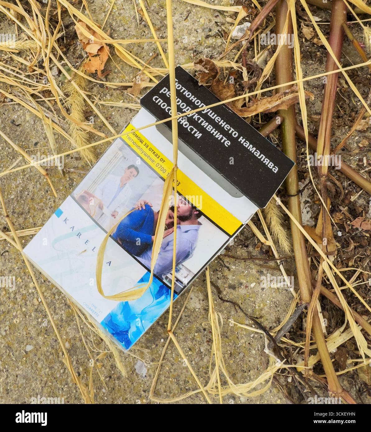 Balchik, Bulgaria - Aug 25, 2025: Close-up of a discarded Karelia cigarette  pack with health warning text in Bulgarian, lying among dry grass and debr  Stock Photo