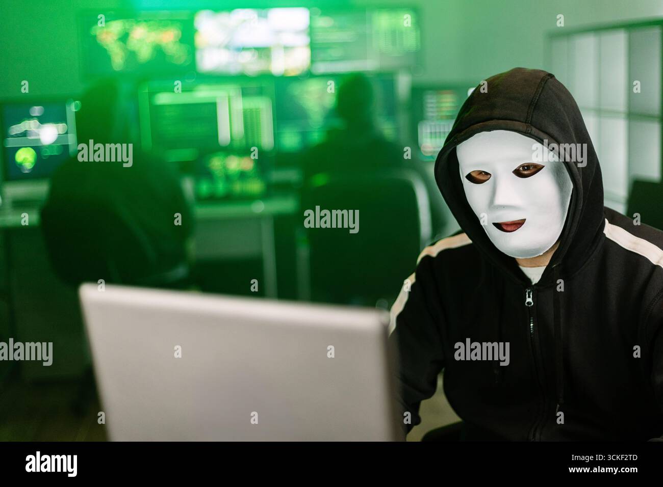 Hacker working on laptop in dark room with green code displayed on monitors Stock Photo