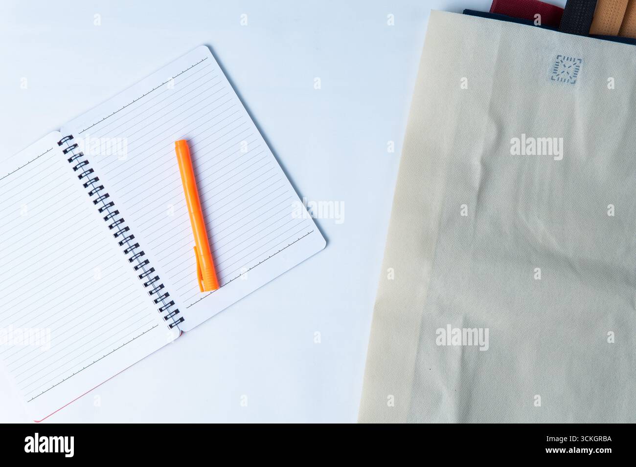 Open spiral notebook with an orange marker placed on lined pages, beside a folded eco bag on a white desk. Stock Photo
