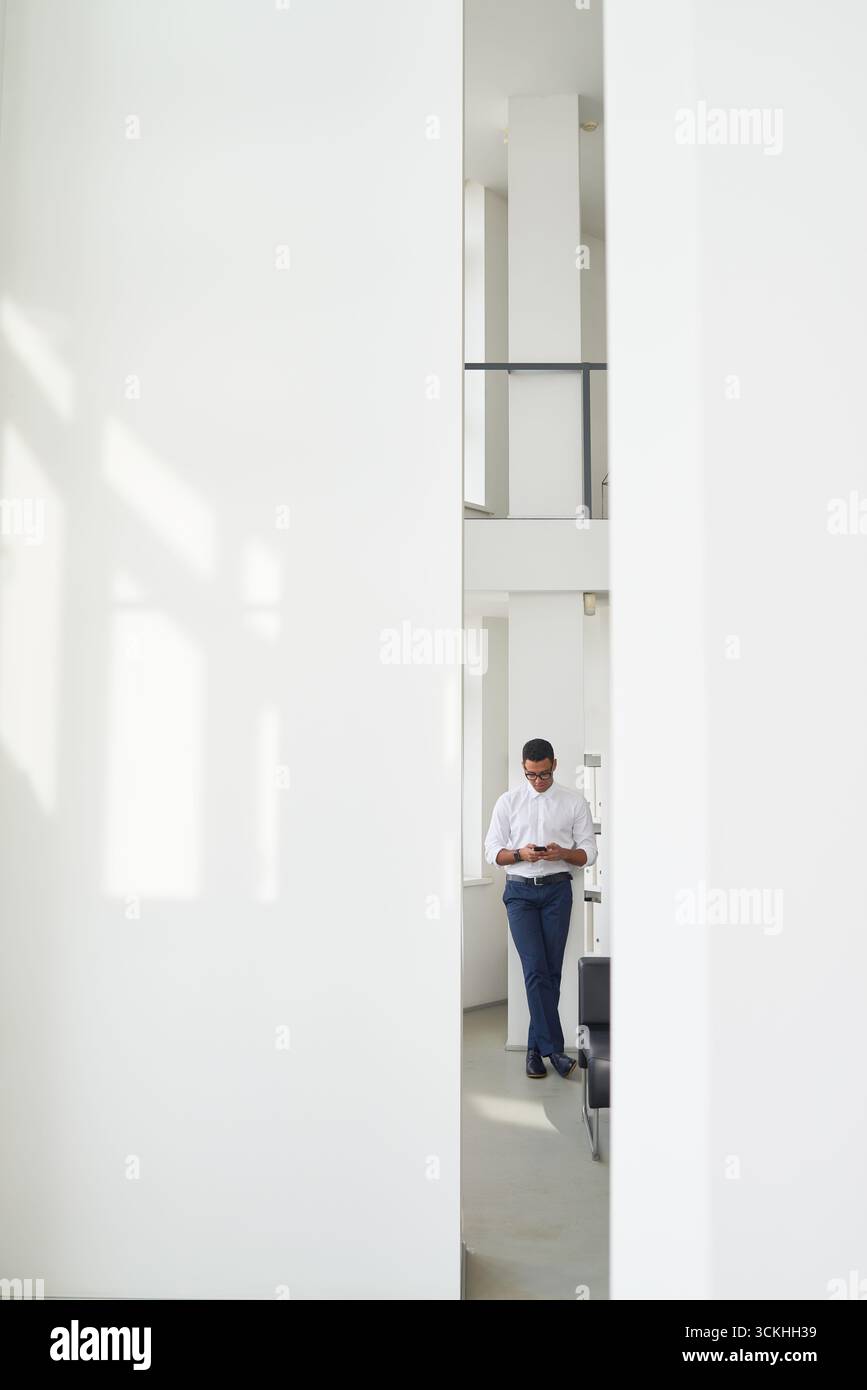Man standing in modern bright office using smartphone, leaning against wall, focused on screen, natural light streaming through large windows Stock Photo