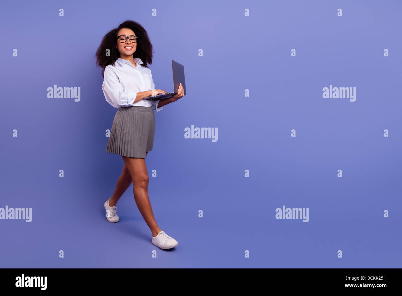 Young professional woman using a laptop while walking against a solid purple background Stock Photo