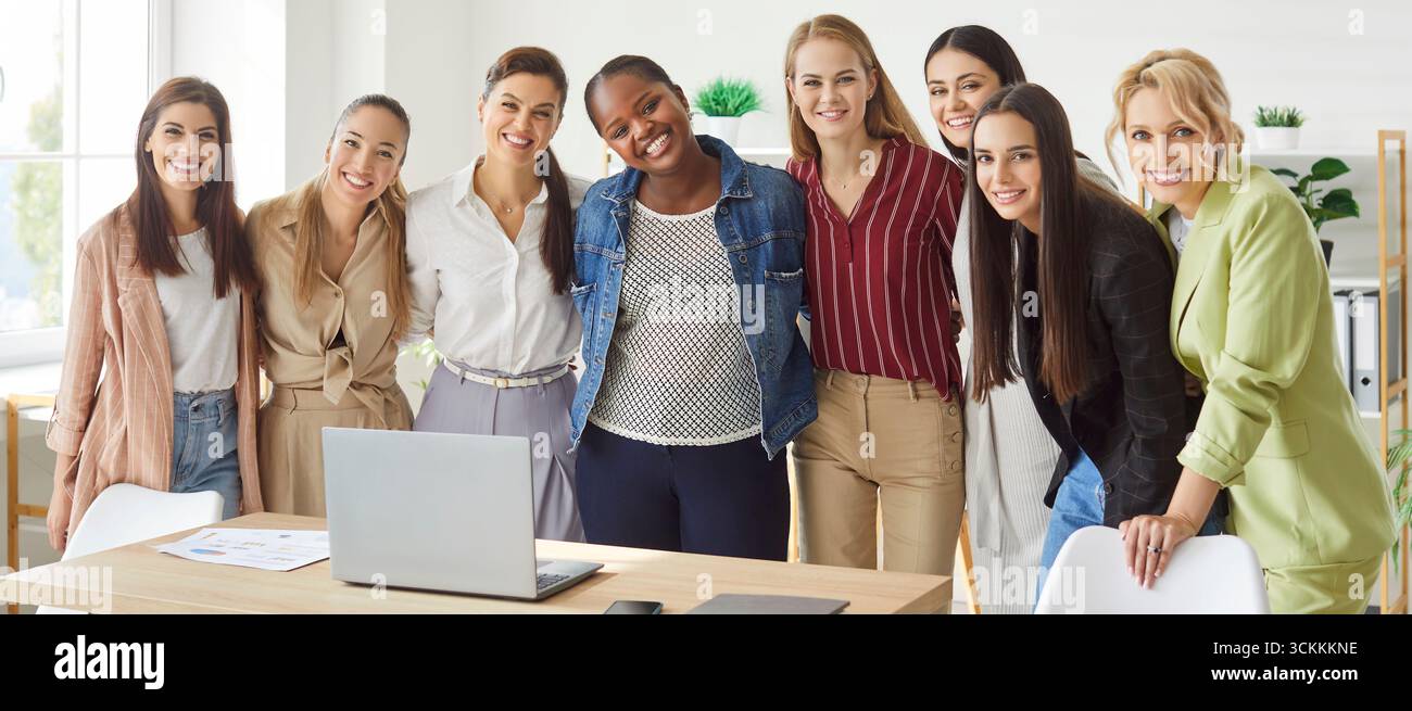 Smiling diverse women standing by desk in office hugging and laughing, showing unity and teamwork. Stock Photo