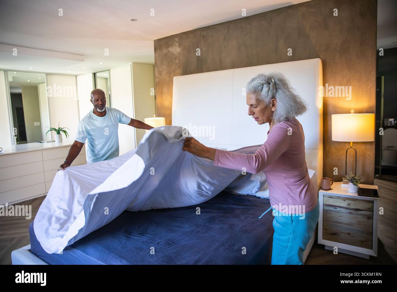 Couple making bed under white headboard in modern bedroom, spreading white bedsheet over mattress Stock Photo