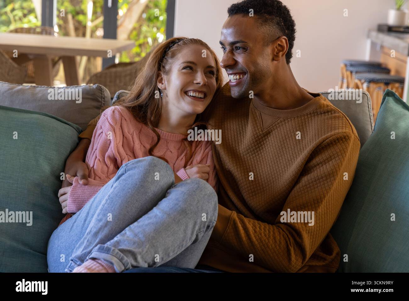 Diverse couple sitting closely on couch in living room by large window and decorative pillows Stock Photo