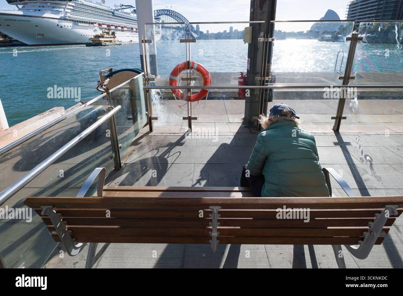 A person sits on a bench under sunlight, facing Sydney Harbour and a docked cruise ship. The serene scene captures the quietude and beauty of maritime Stock Photo