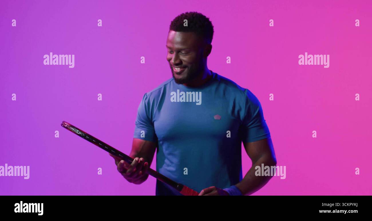 Smiling man wearing blue tee and wristband inspecting black racket under magenta lights in studio Stock Photo