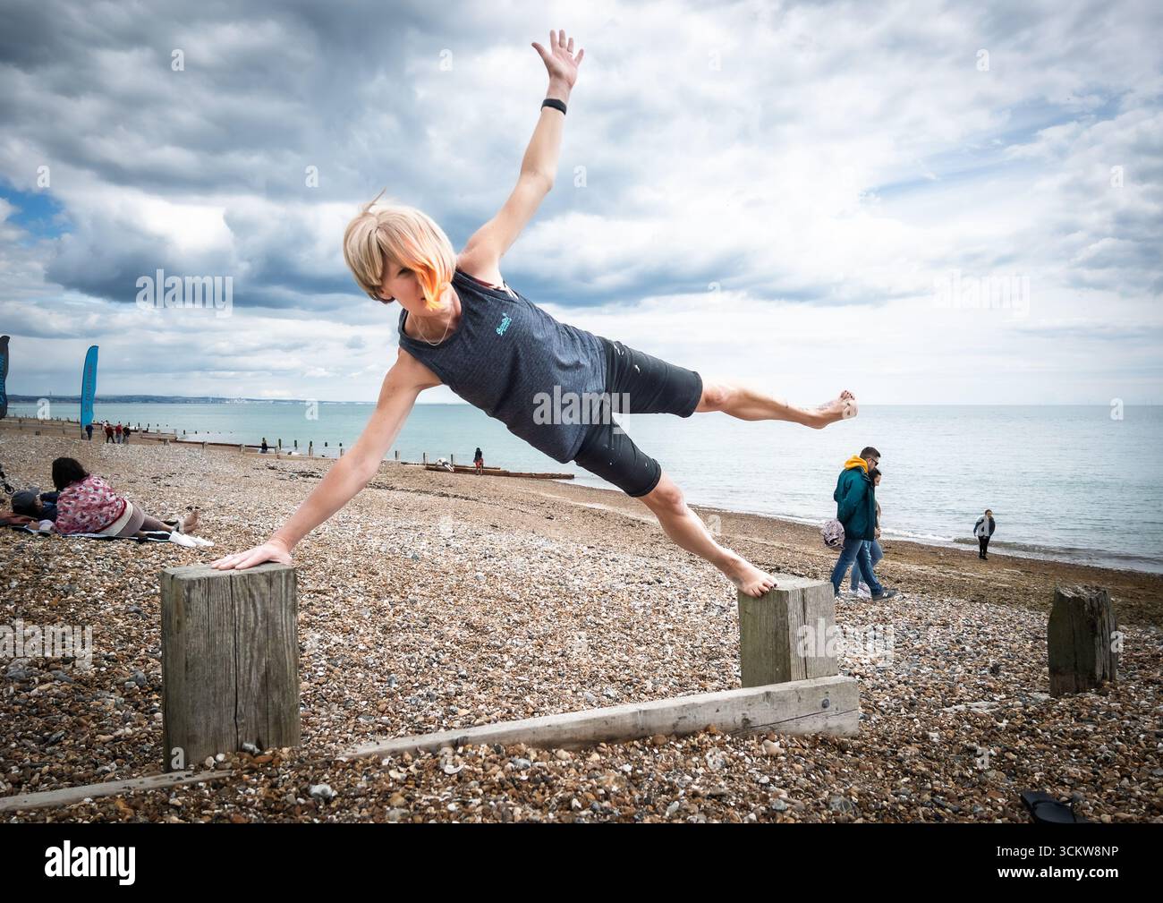 A woman doing a yoga side plank pose on the beach, balancing on the wooden posts of a groyne - Smartphone Captured Stock Image