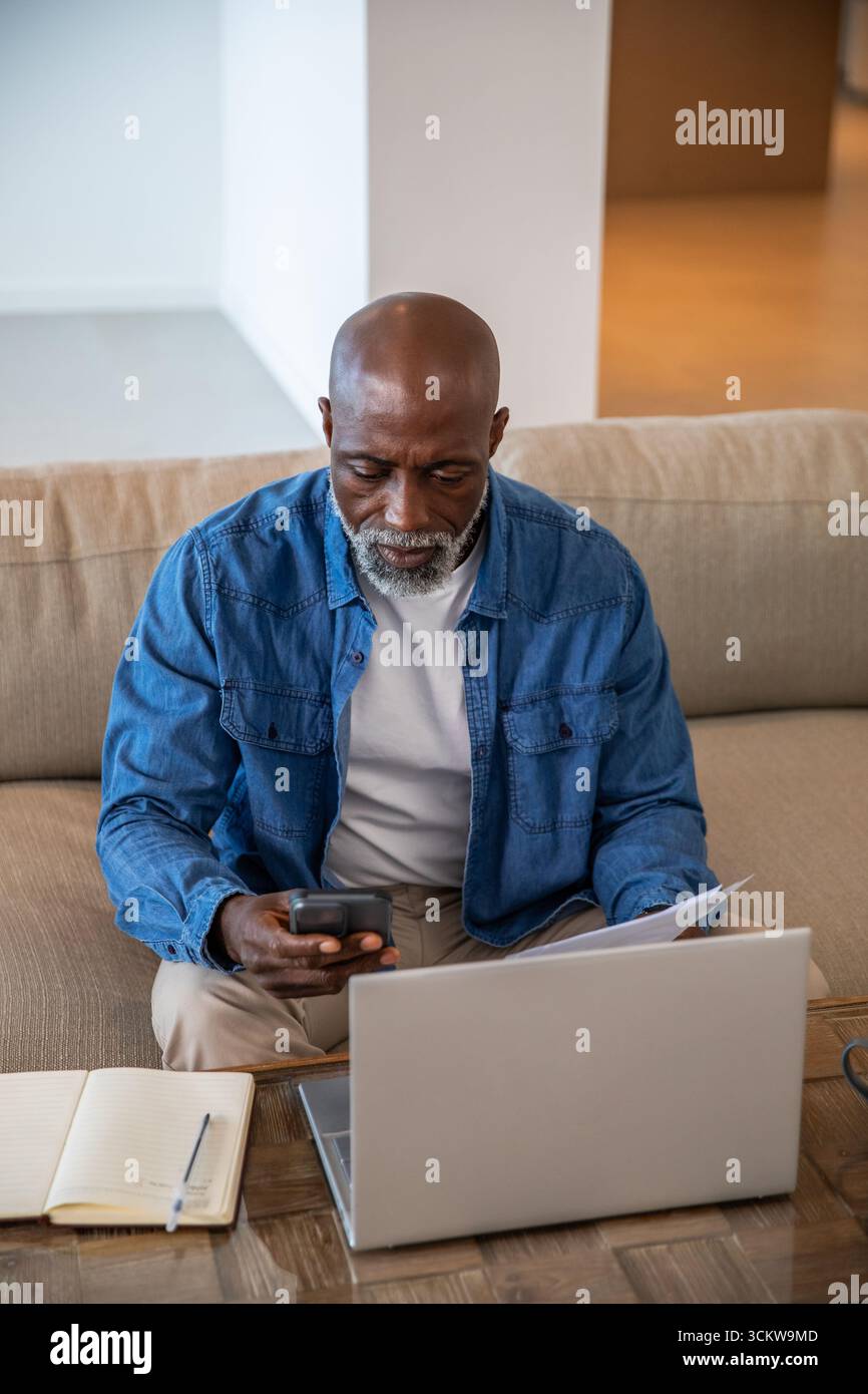 African american senior man using laptop and smartphone, reviewing papers and notebook at home Stock Photo