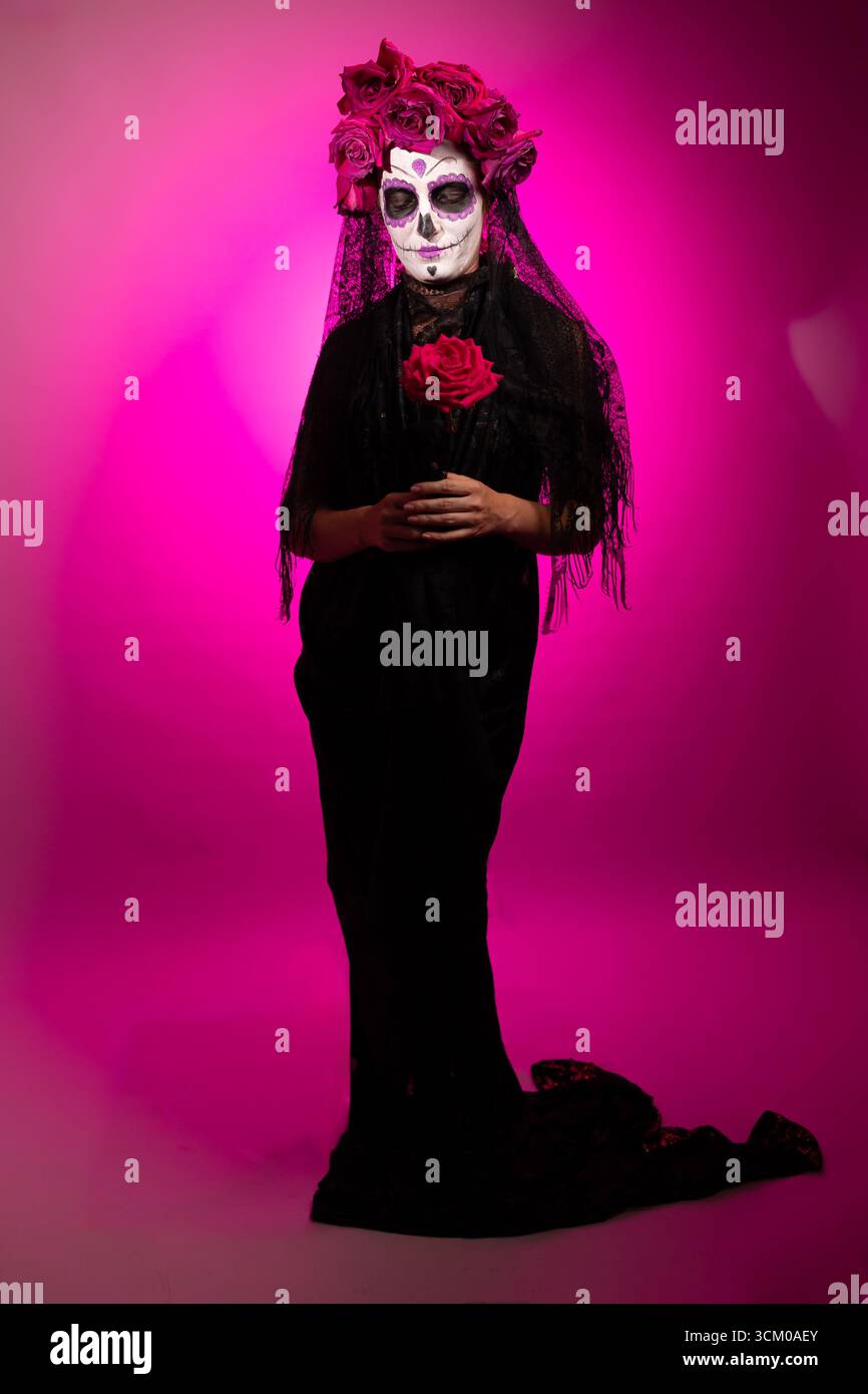 Woman dressed as a Calavera Catalina is standing against a magenta background. She wears sugar skull makeup and a flower crown and holds a red rose in her hands. Stock Photo