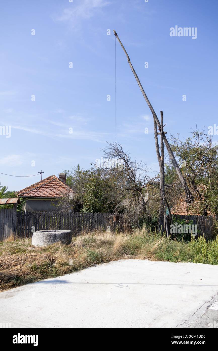 Old rural water well with a wooden pole lifting system in front of a countryside house, surrounded by dry grass and trees under a clear blue sky Stock Photo