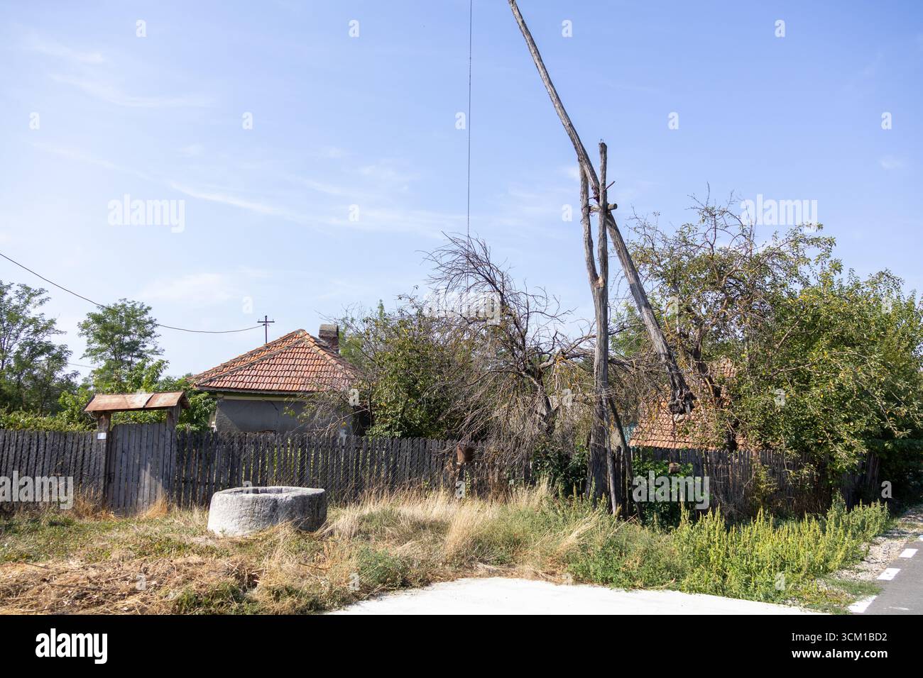 Old rural water well with a wooden pole lifting system in front of a countryside house, surrounded by dry grass and trees under a clear blue sky Stock Photo
