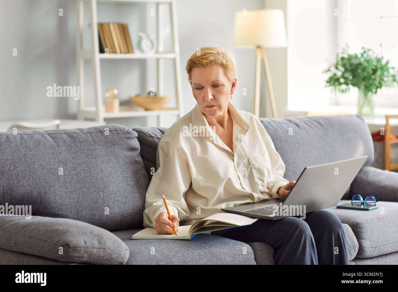 Mature woman is focused on work, taking notes in notebook while using her laptop. Stock Photo