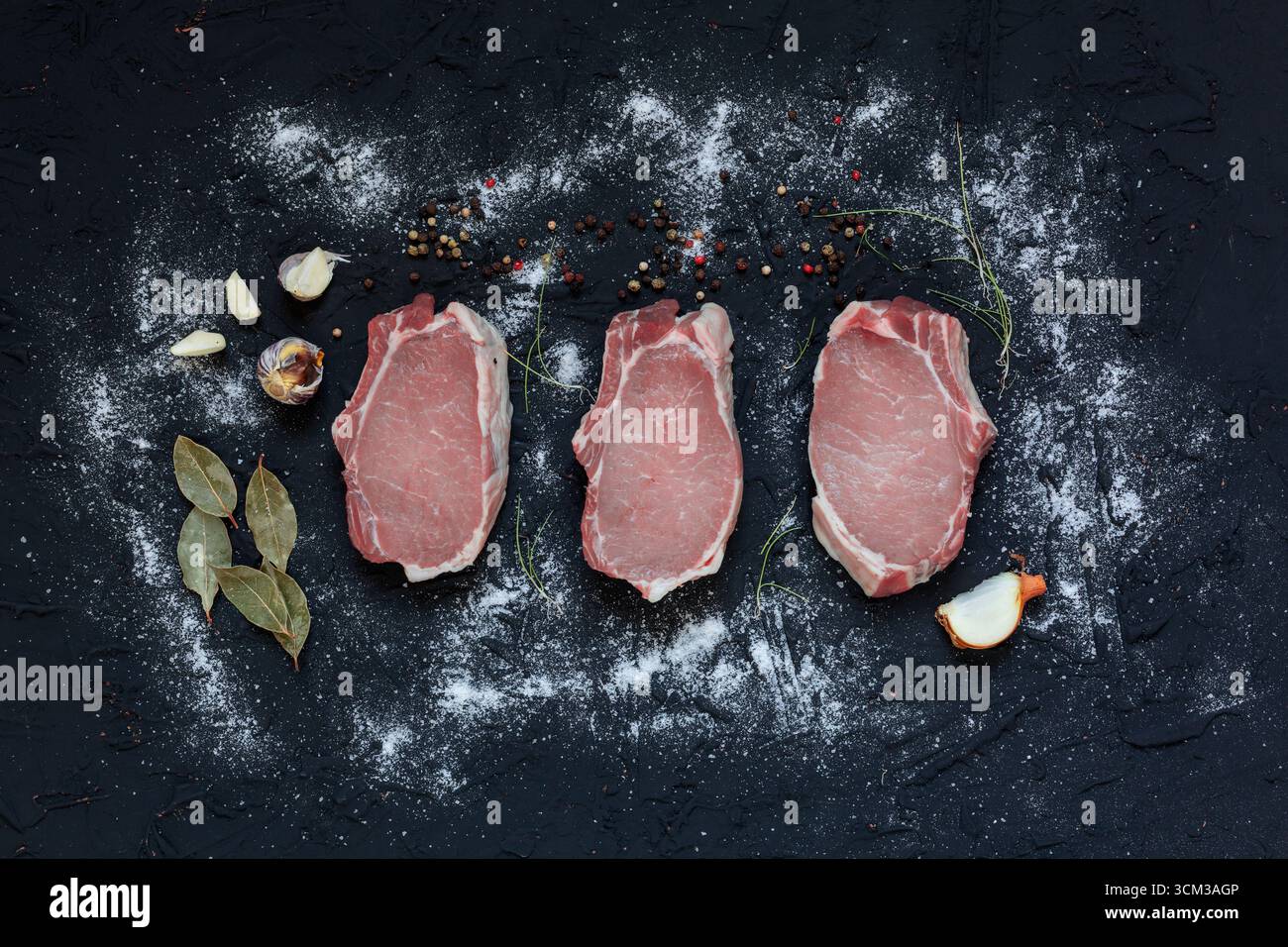 Top view of three fresh raw pork chops placed on a dark textured surface with scattered flour, peppercorns, garlic cloves, bay leaves, onion, and herb Stock Photo