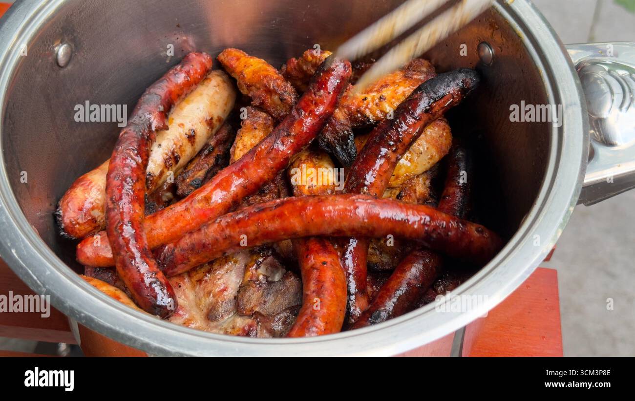 A close-up overhead view of a pot filled with various grilled meats, primarily sausages and chicken wings, with tongs reaching in. Stock Photo
