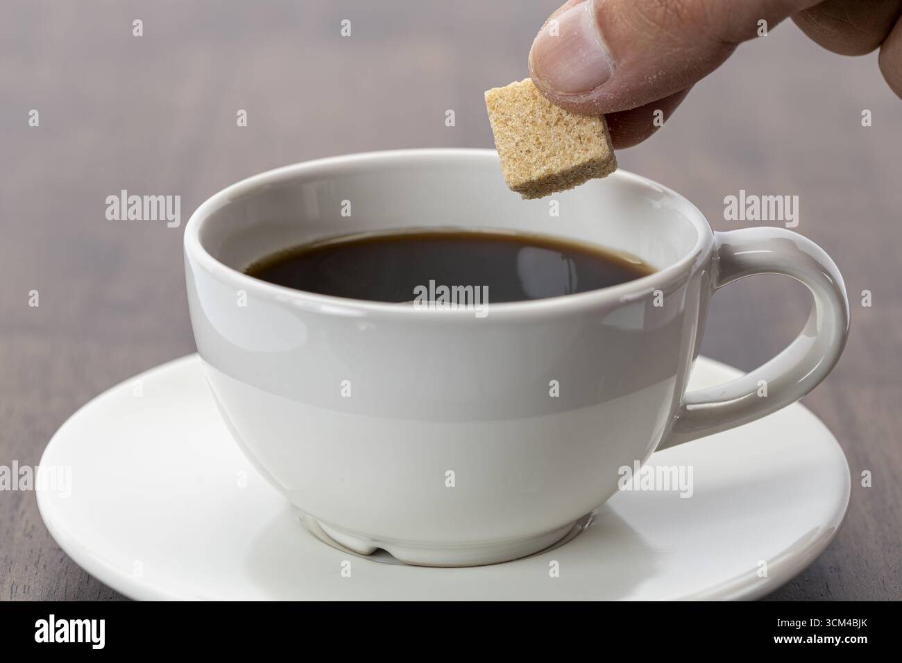 A hand holding organic sugar cube above hot black coffee in white ceramic cup on saucer, preparing to sweeten drink, morning beverage concept, close u Stock Photo
