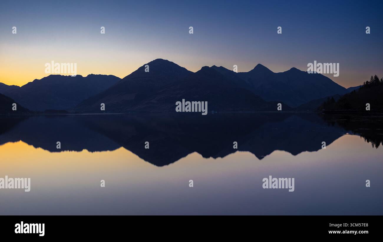 View across Loch Duich of the silhouette of the Five Sisters mountain range at sunrise. The mountains are reflected in the water. Stock Photo