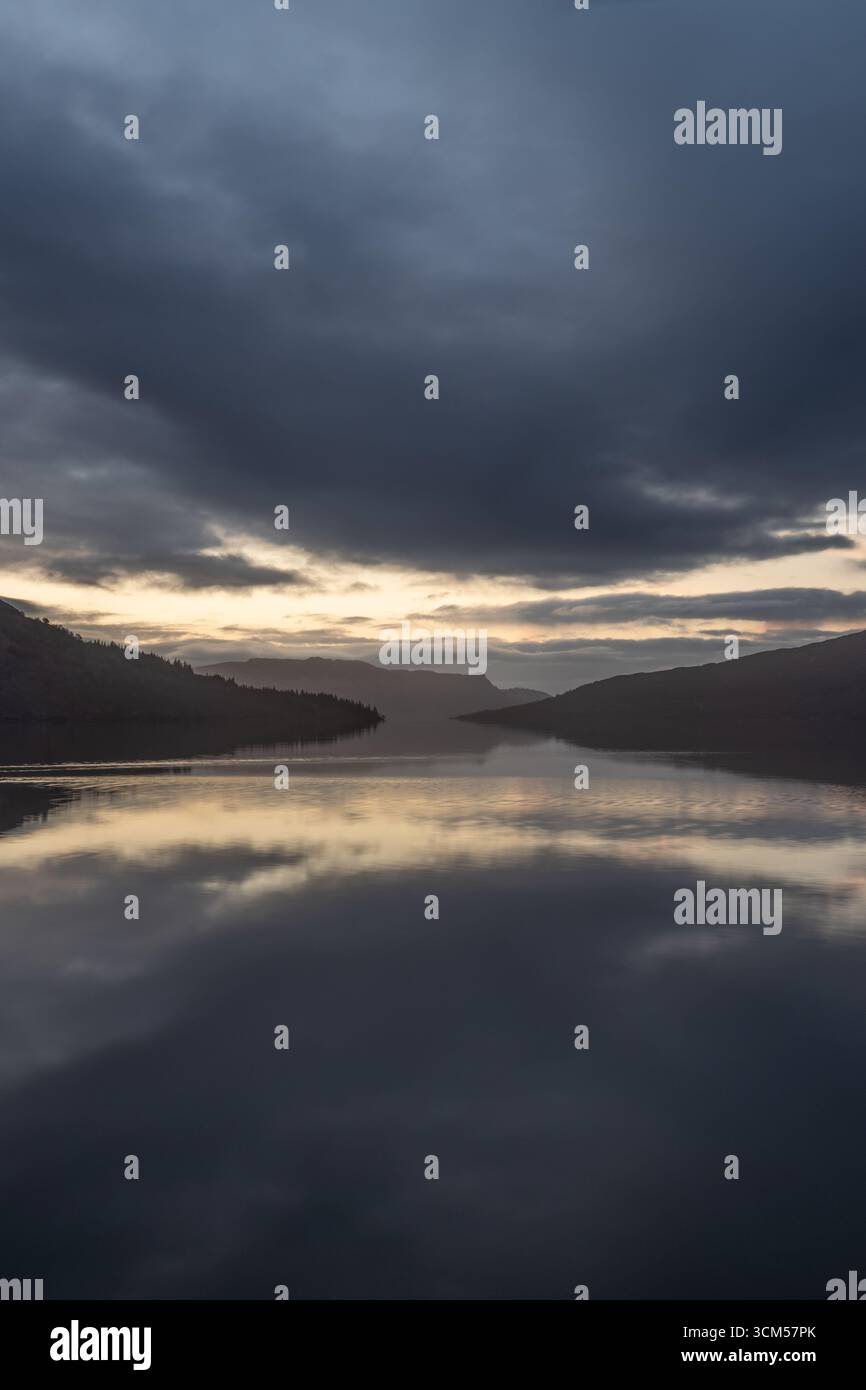 Blue hour with a gap in the clouds on the shore of Loch Katrine. The clouds and mountains are reflected in the water. Stock Photo