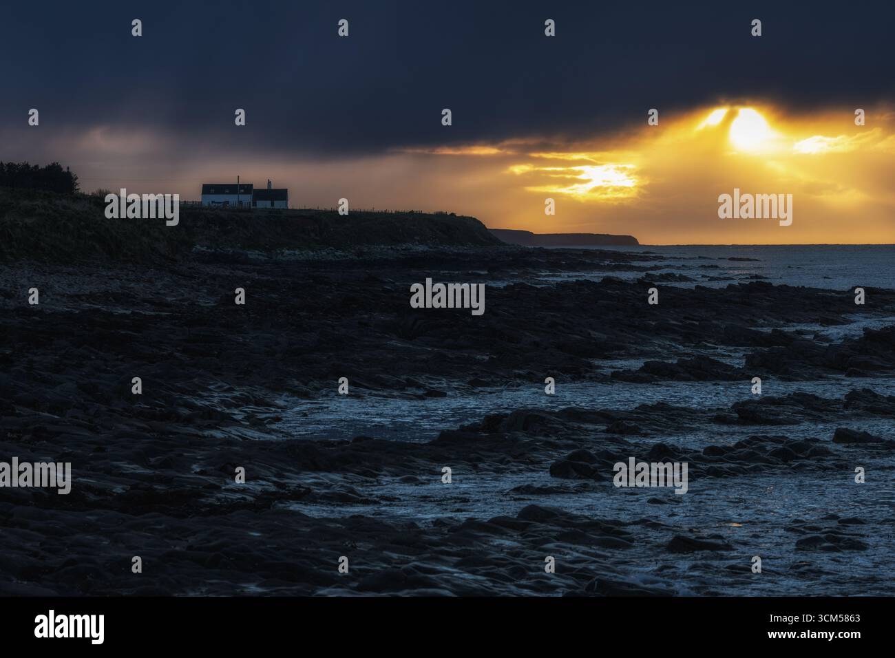 A lonely house stands on the coast. The sky is dark and threatening. Yellow sunlight shines through a gap in the clouds. Stock Photo
